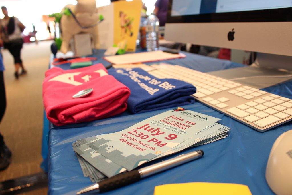 Photo of the BIG IDeA display booth at the 2017 Maker Festival. Table with bright pink and blue team shirts, and brochures on it.