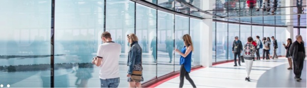 This image is of a photograph that shows a wall of glass that stretches from floor to ceiling, with people standing around looking out. Out the windows, you can see some of Toronto Island and Lake Ontario.