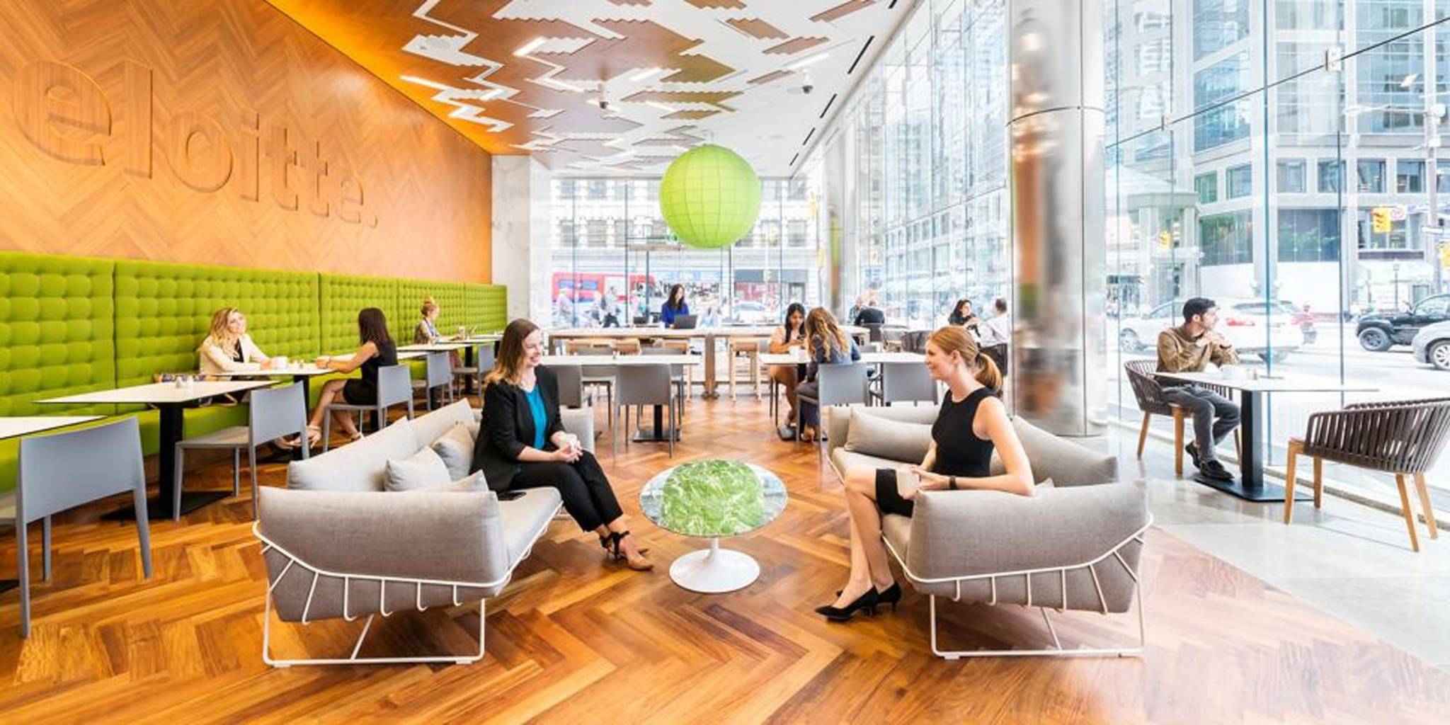 This image is a photograph of the inside of the Deloitte building in a cafe style room and includes people sitting down some of whom are socializing.