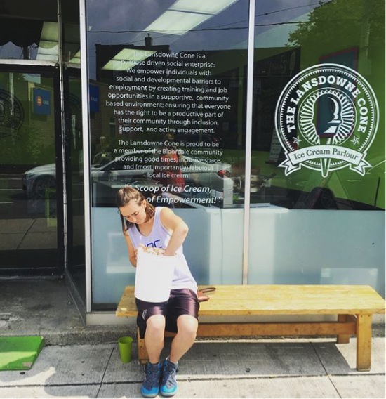 This image is of a photograph that shows someone sitting on a bench outside the Lansdowne Scoop, scooping ice cream from a very large bucket. Visible in the photo is a small green ramp from Stopgap.ca. Also visible on the storefront glass is a message from the store that reads: “The Lansdowne Cone is a purpose-driven social enterprise. We empower individuals with social and developmental barriers to employment by creating training and job opportunities in a supportive, community-based environment; ensuring that everyone has the right to be a productive part of their community through inclusion, support and active engagement. The Lansdowne Cone is proud to be a member of the Bloordale community providing good times, inclusive space and (most importantly) fabulous local ice cream! A Scoop of Ice Cream, and a Heap of Empowerment.”