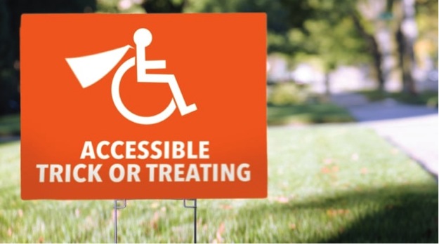 This image is of a photograph that shows an orange coloured sign planted on a lawn of grass. The sign features an image of the international symbol of access with a superhero cape added to the design.