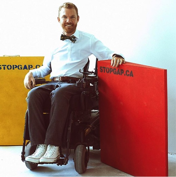 This image is a photograph of Luke Anderson, using a power wheelchair, sitting in between 2 wooden wedge ramps that are standing upright on their sides, one is painted yellow the other painted red. The ramps read stopgap.ca on them.
