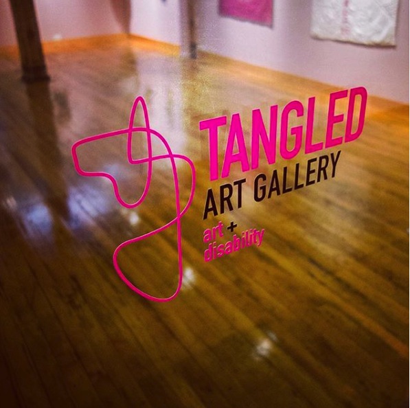 Image is a photograph of the gallery logo and name “Tangled Art Gallery art + disability” pasted on the glass doors of the gallery. Seen beyond the logo is the actual gallery space showing an open space, as well as art hung on the wall at a lower height than what is seen in most other galleries. This is done to accommodate people in a seated position.