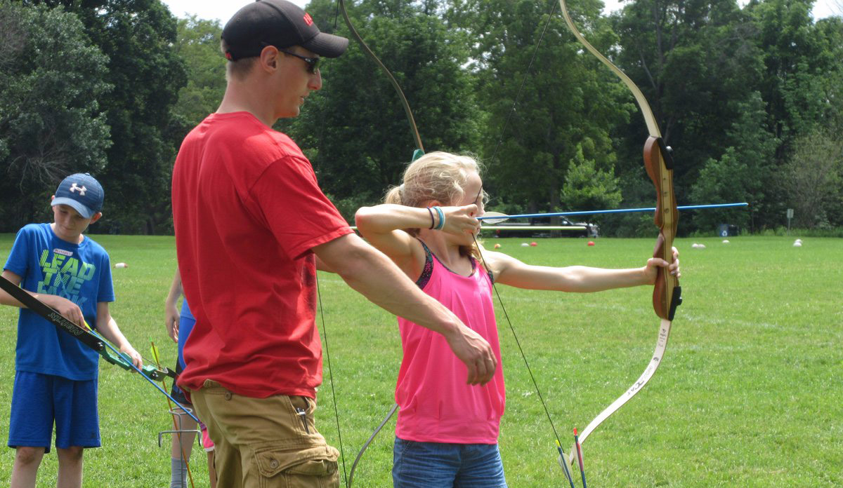 This image is a photograph that shows an instructor teaching a little blond girl archery.