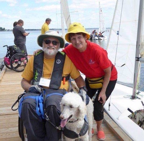 This image is a photograph showing a wheelchair user and their service dog. Next to the wheelchair user is someone with their arm around them. They are all beside a sailboat on a dock. In the background is an empty wheelchair, implying that someone is already in a boat, sailing.